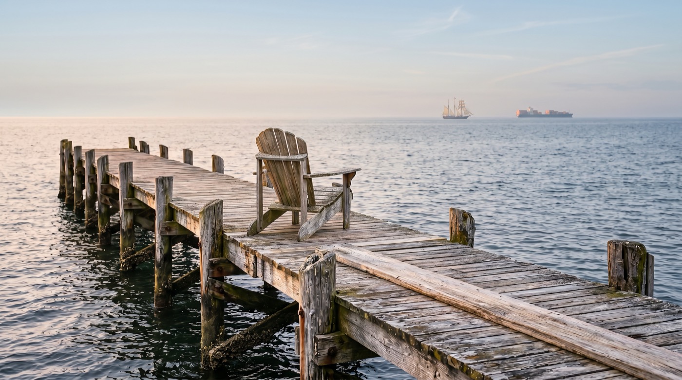An empty Adirondack chair at the end of a weathered wooden pier, looking out over calm water toward a tall ship and a container ship on the horizon