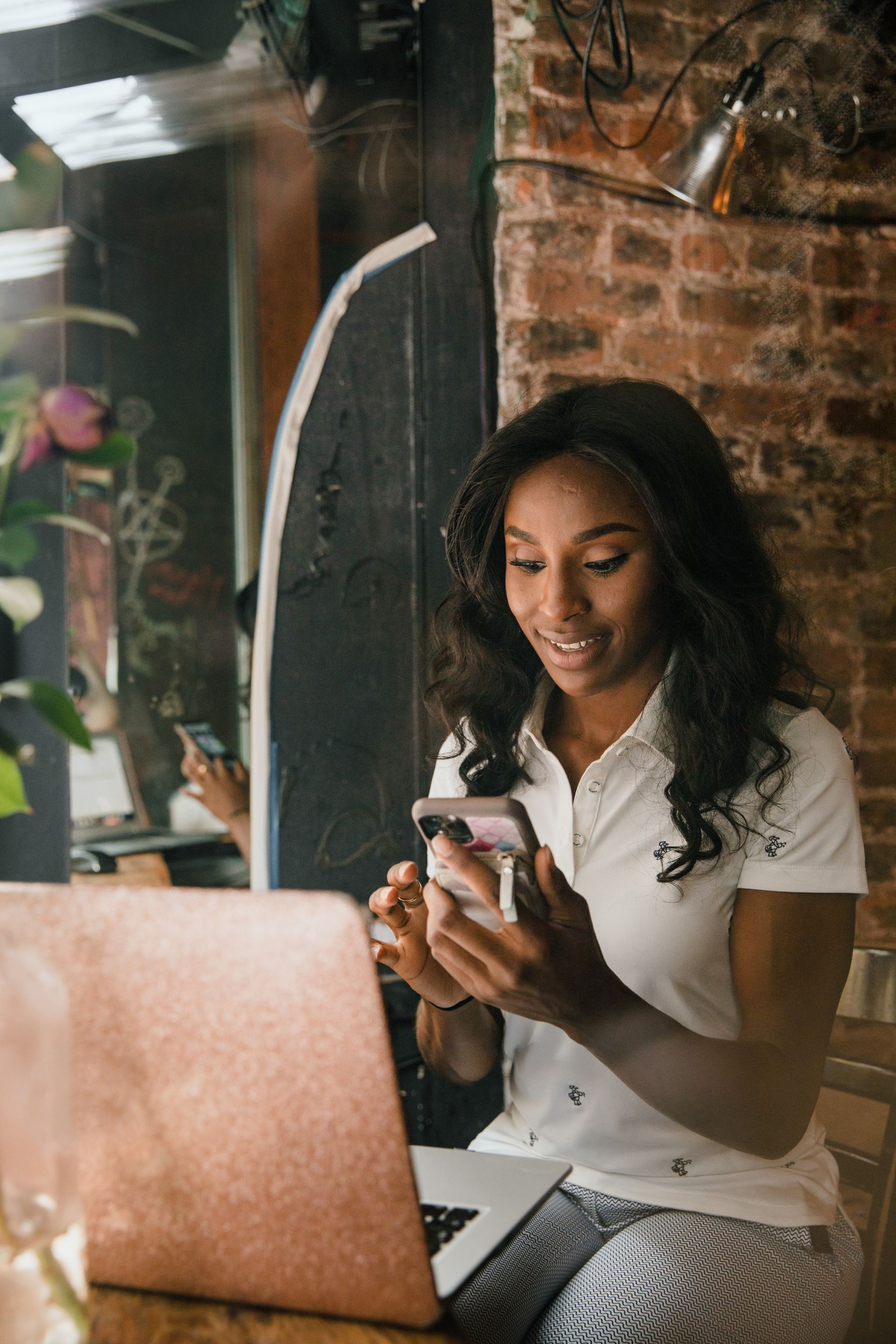 A small-business owner smiling as she reads a message on her phone at her café counter, a laptop open in front of her